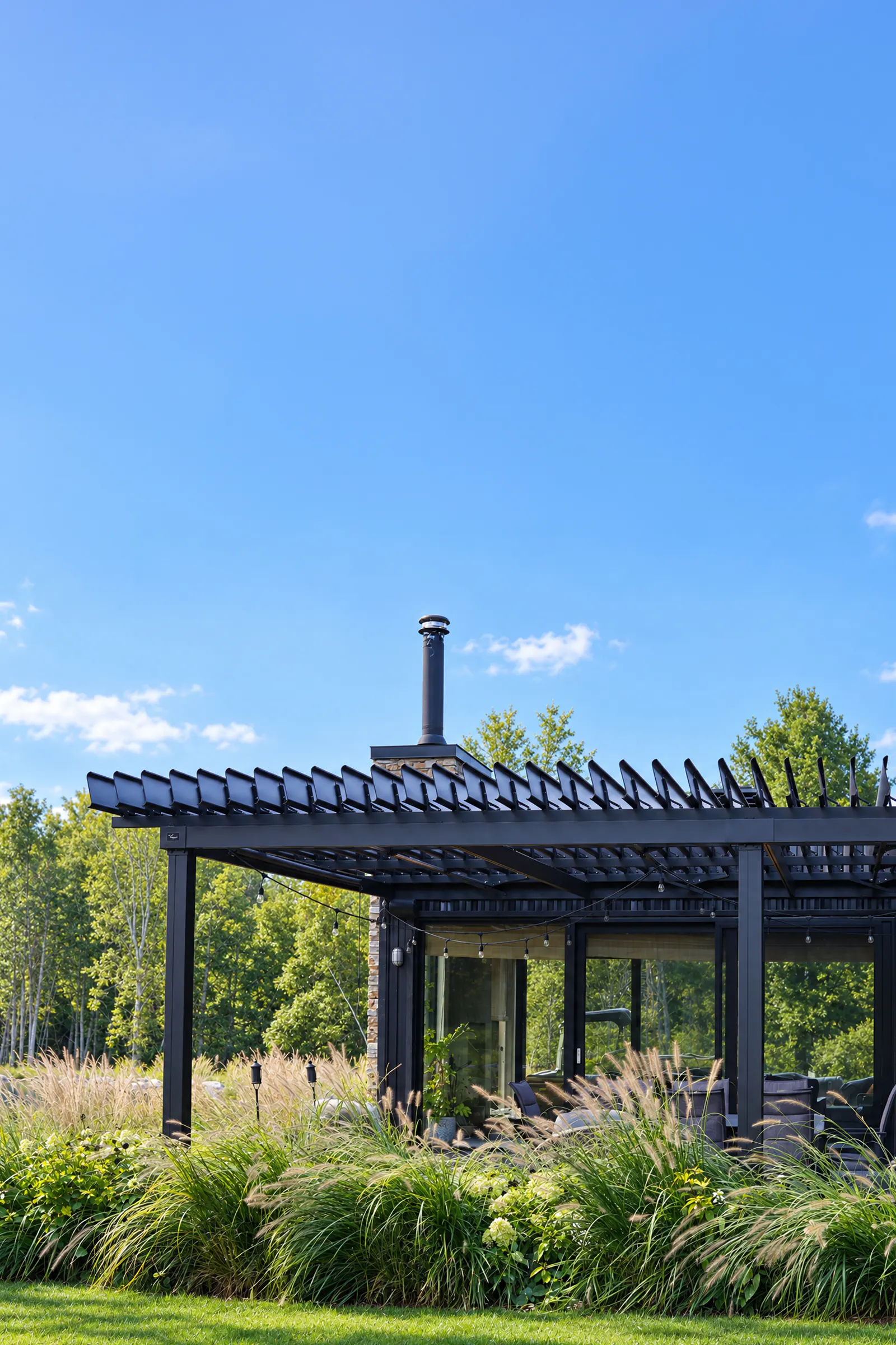 Pergola SunLouvre Pergolas noire à lames orientables au-dessus d'une terrasse, entourée de graminées, sous un ciel bleu