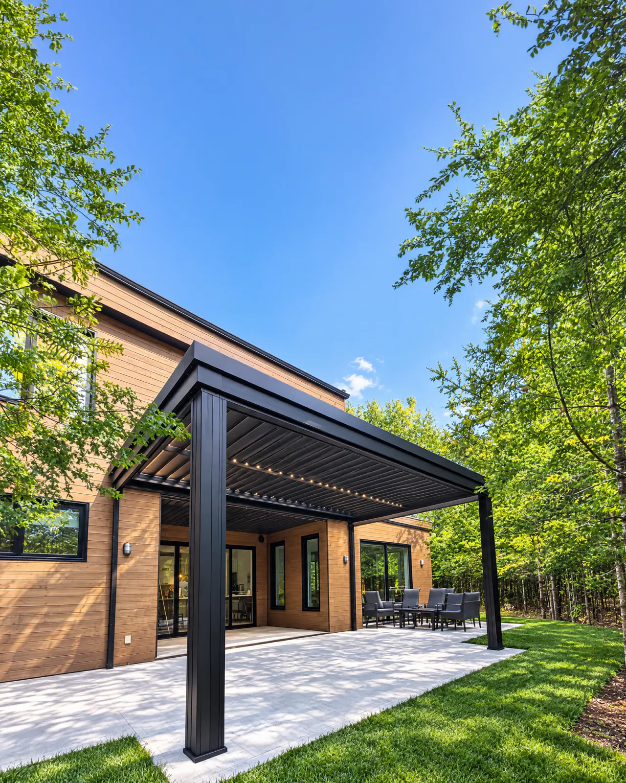 Pergola à lames intégrées noire avec éclairage DEL, le long d'une maison au revêtement de bois, au-dessus d'un patio