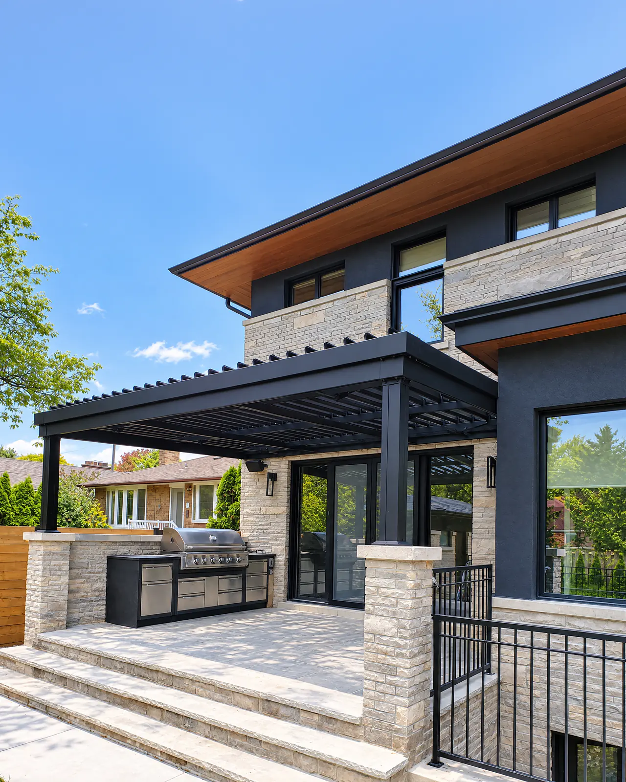 Pergola à lames intégrées noire attachée à une maison en pierre, au-dessus d'une terrasse avec cuisine extérieure