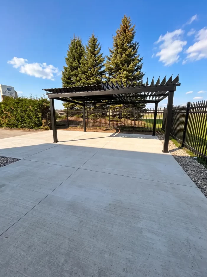 Black pergola on a concrete pad near the conifers, Fempro in Drummondville