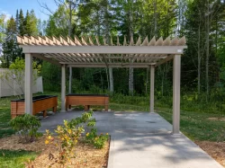 Pergola beige au-dessus d'une aire pavée avec bancs, en lisière de bois, Maison Catherine de Longpré à Saint-Georges-de-Beauce